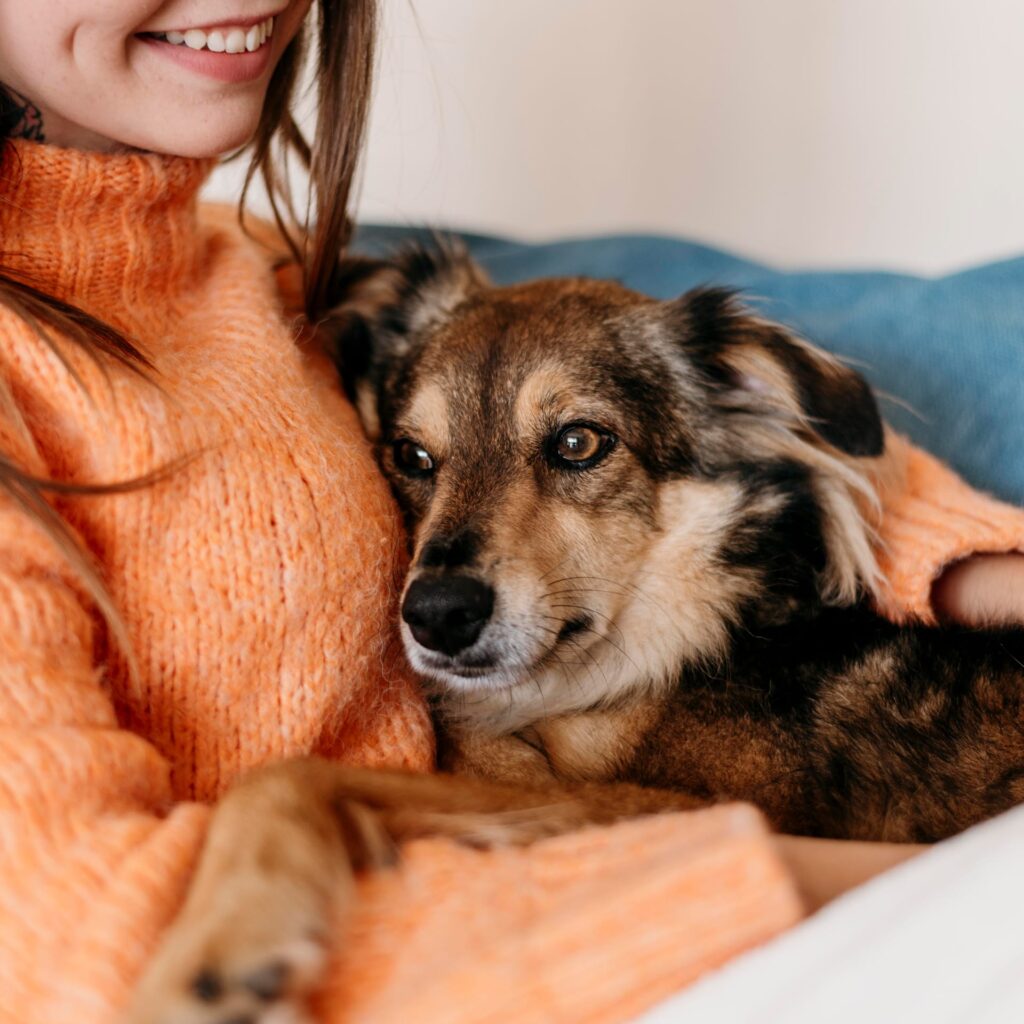 O femeie zâmbitoare într-un pulover portocaliu îmbrățișează un câine cu blană maro și bej