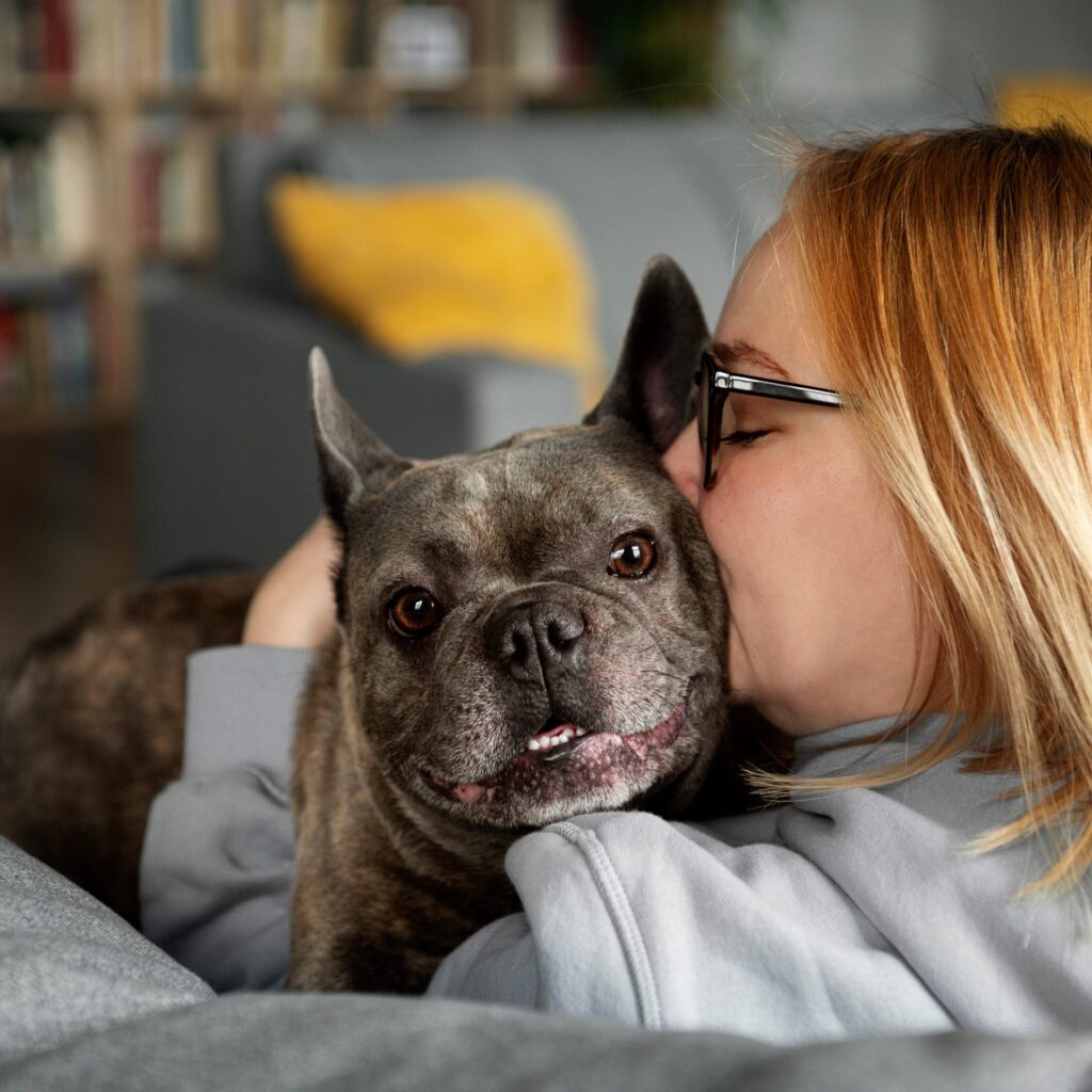 O femeie cu ochelari sărută afectuos un buldog francez pe cap, stând pe canapea